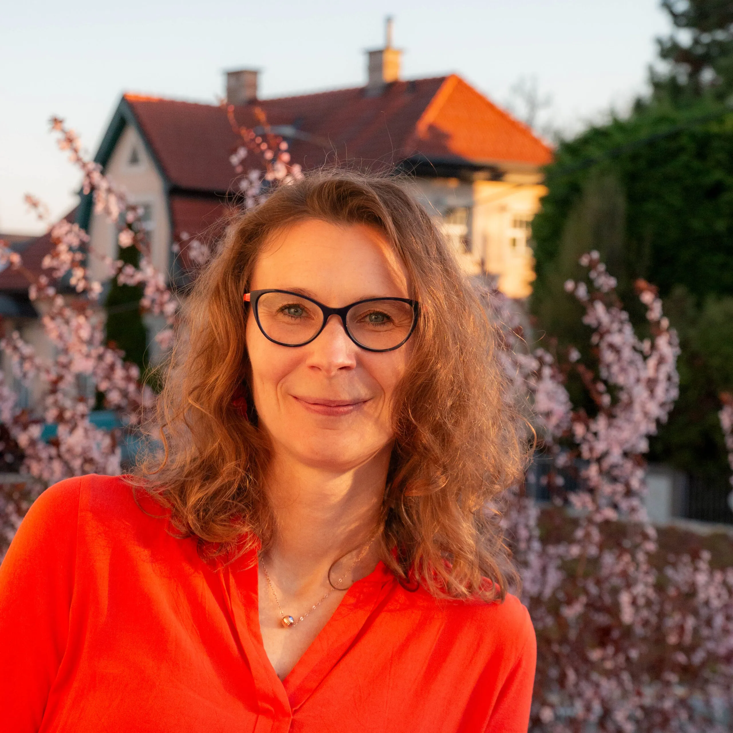Ein Porträtfoto von Martina Lichtblau in der Natur. Hinter ihr blüht ein Baum mit rosa Blüten. Sie trägt einen orangefarbenen Pullover und sieht freundlich aus.