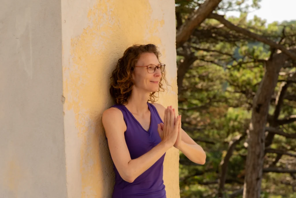 Martina Lichtblau lehnt an einer Wand in der Natur und macht mit ihren Händen eine dankbare Meditationsgeste. Sie sieht glücklich und zufrieden aus.