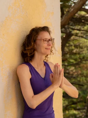 Martina Lichtblau lehnt an einer Wand in der Natur und macht mit ihren Händen eine dankbare Meditationsgeste. Sie sieht glücklich und zufrieden aus.